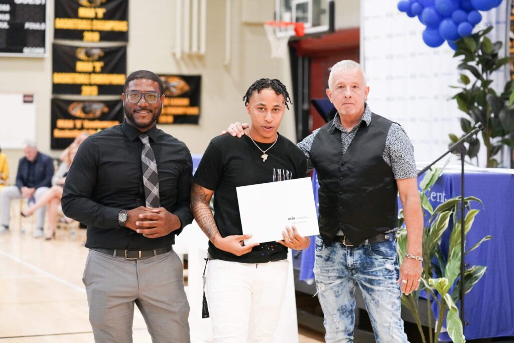 3 men standing at NCIA graduation, 1 holds a certificate