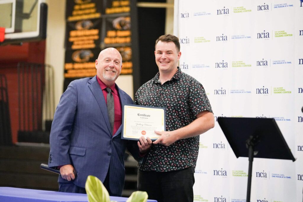 two men standing at graduation, one is holding a certificate from NCIA