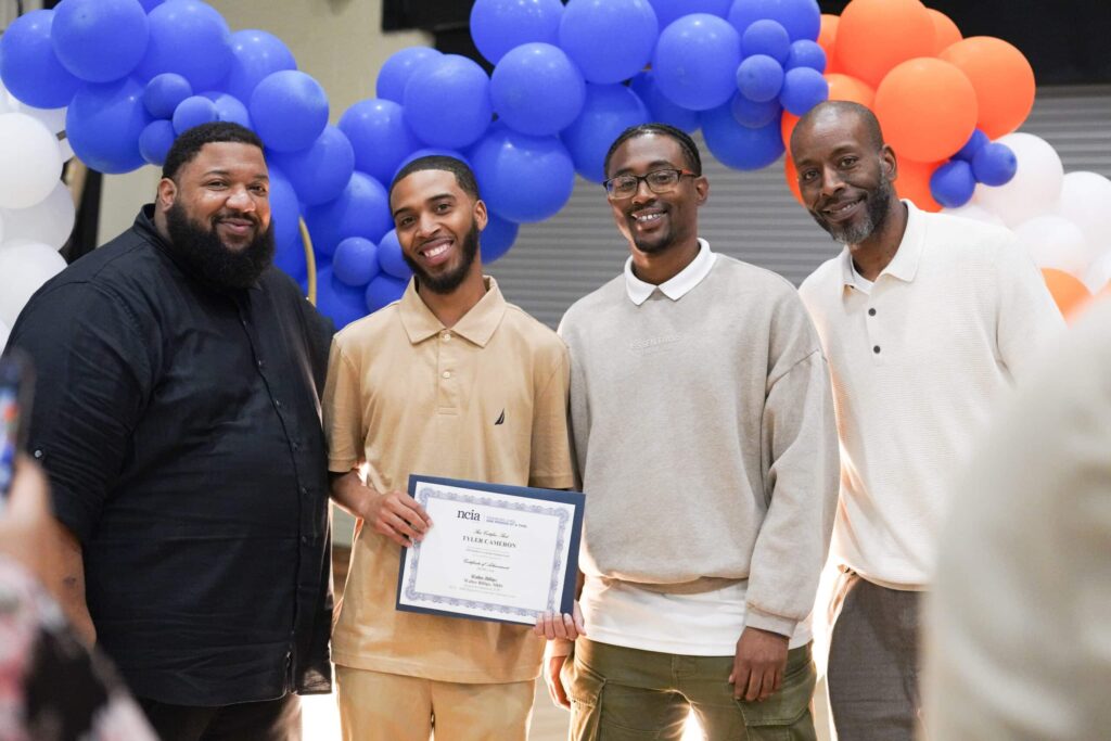 4 men standing at graduation, 1 is holding a certificate